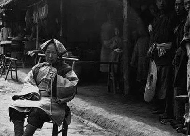 An elderly woman with bound feet sitting and smoking in front of a door.