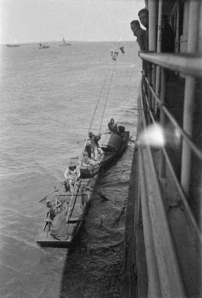 A beggar on a wooden boat, reaching out for alms from those on a ship.