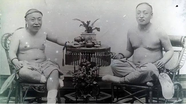 Two shirtless men sit enjoying tea.