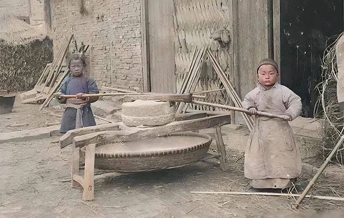 Two boys around six or seven years old pulling a millstone in front of their house.