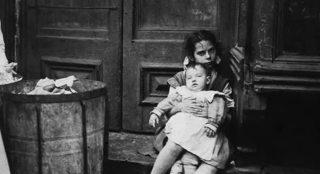 A young child holding a baby sitting outside a home in New York City around 1890