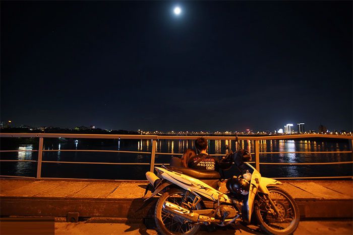 A young couple stops on Long Bien Bridge, sitting together to admire the full moon