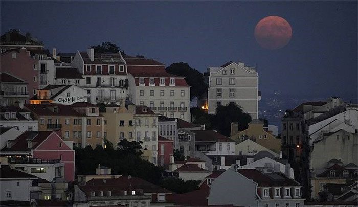 The silhouette of a red supermoon appearing hazy behind the Castelo neighborhood of Lisbon.